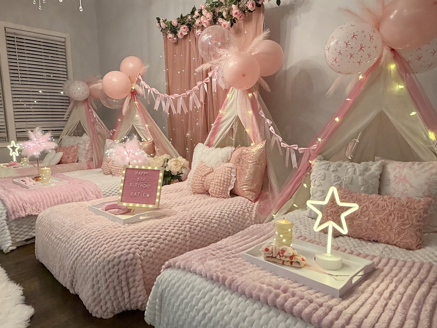 Coquette sleepover setup with a rose garland backdrop, bow-print balloons, ribbon banners and blush rosette pillows