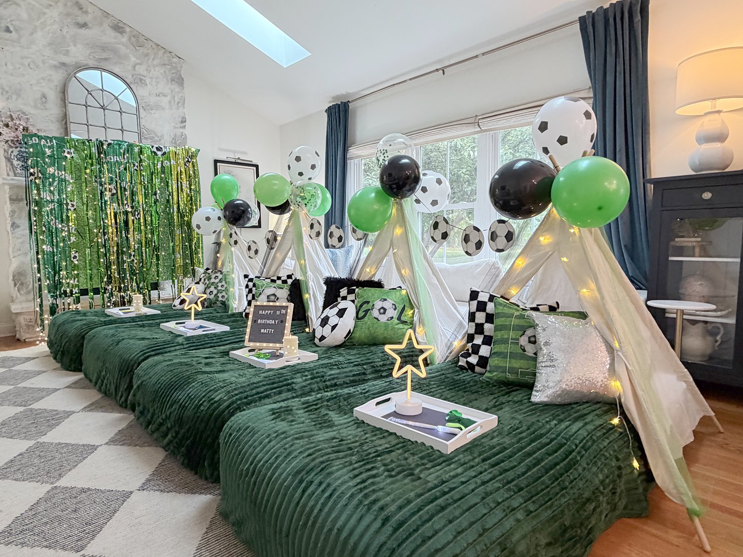 Soccer sleepover setup with green fringe backdrop, soccer ball garland, checkered pillows and deep green bedding